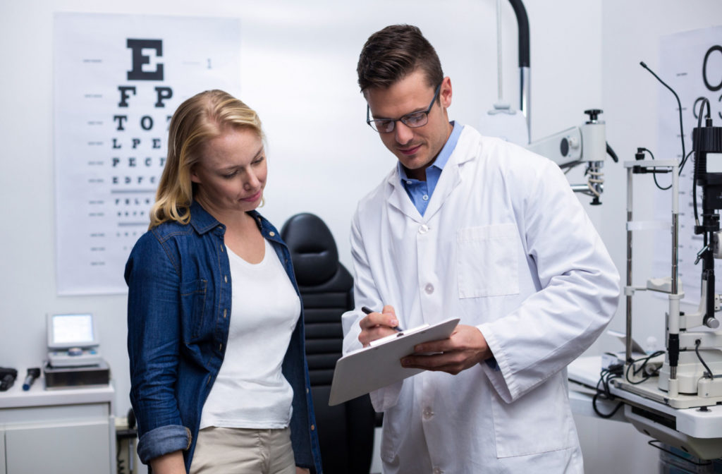 A male doctor holding a medical chart and a ballpen that is standing beside a female patient discussing the information in the chart.