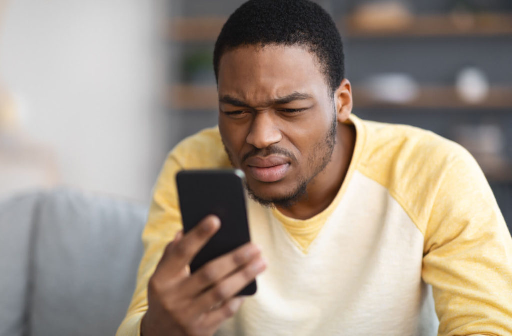 A man sitting on a sofa is squinting his eyes while using his smartphone. Squinting is one of the signs of light sensitivity.