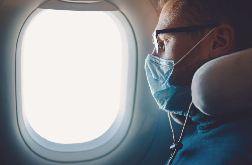 a man sitting on a plane with a face mask on, looking out the airplane window